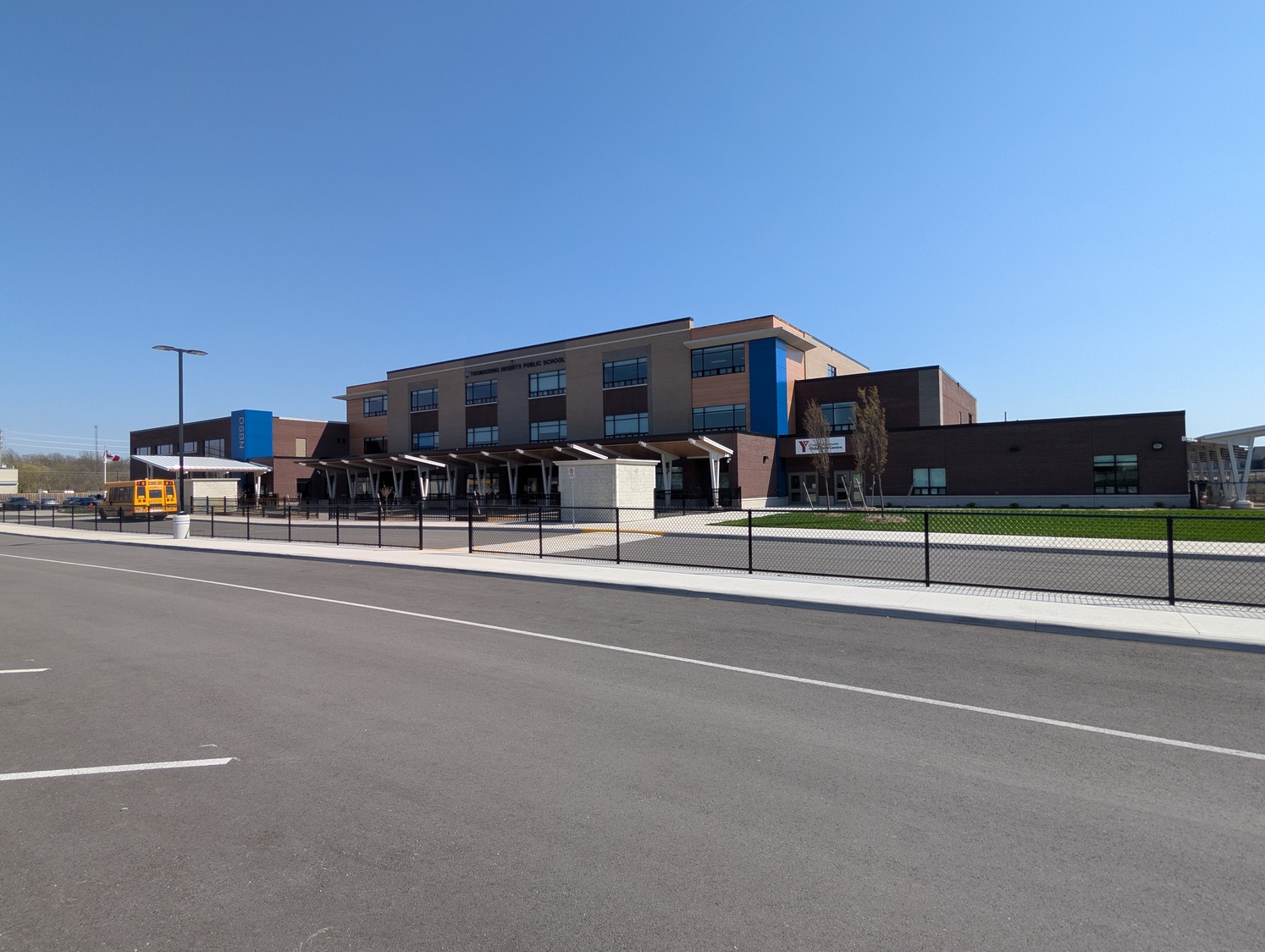 A modern, three-story Thundering Heights school building with large windows, a fenced lawn, and a yellow school bus parked outside under a clear blue sky.