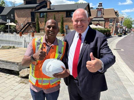 Two men, one in a safety vest holding a hard hat and the other in a suit, smile and give thumbs up on a sunny street with brick buildings in the background.