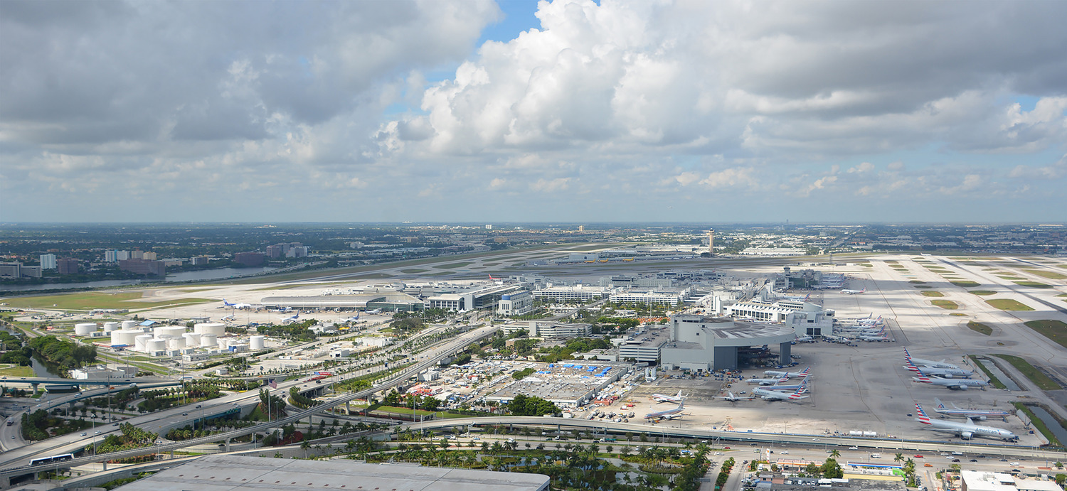 Aerial view of a large airport with multiple runways, taxiways, terminal buildings, parked airplanes, and fuel storage tanks under a partly cloudy sky showcases modern airport design.
