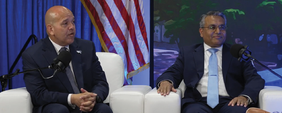 Two men in suits sit on white chairs, speaking into microphones, at CoMotion Miami 2026. An American flag and a painting are visible in the background.