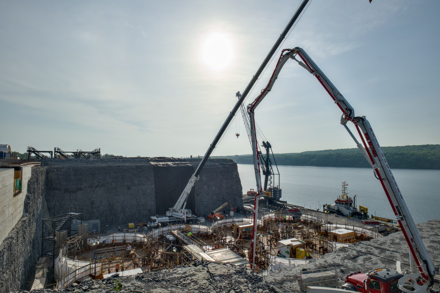 Construction site beside a body of water with cranes and equipment, concrete forms, and workers under a clear sky with the sun overhead.