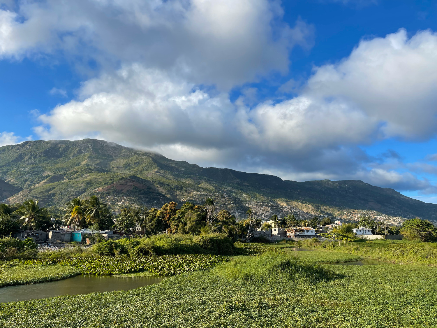 A lush green field and river in the foreground, with small houses scattered below a mountain range under a partly cloudy blue sky.