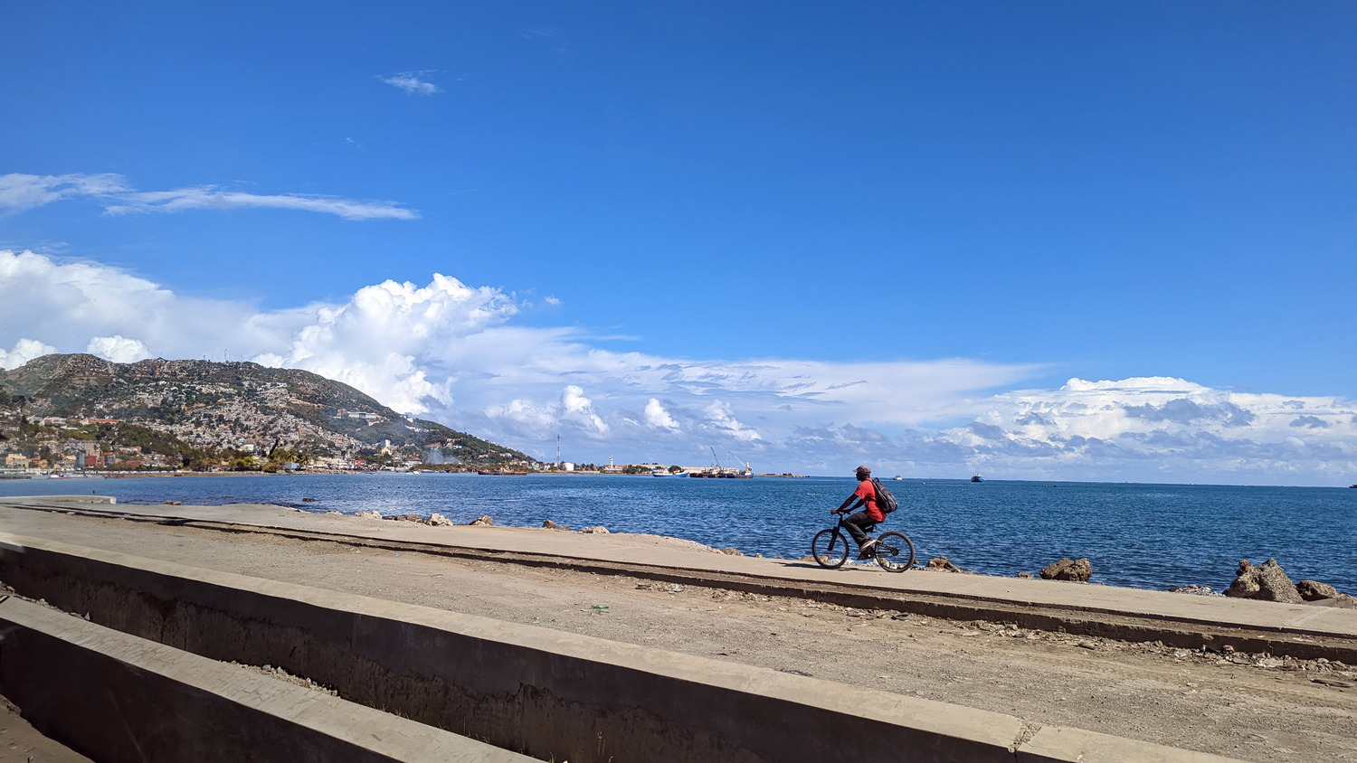 A person rides a bicycle along a coastal path with the sea on one side and mountains in the background under a partly cloudy sky.