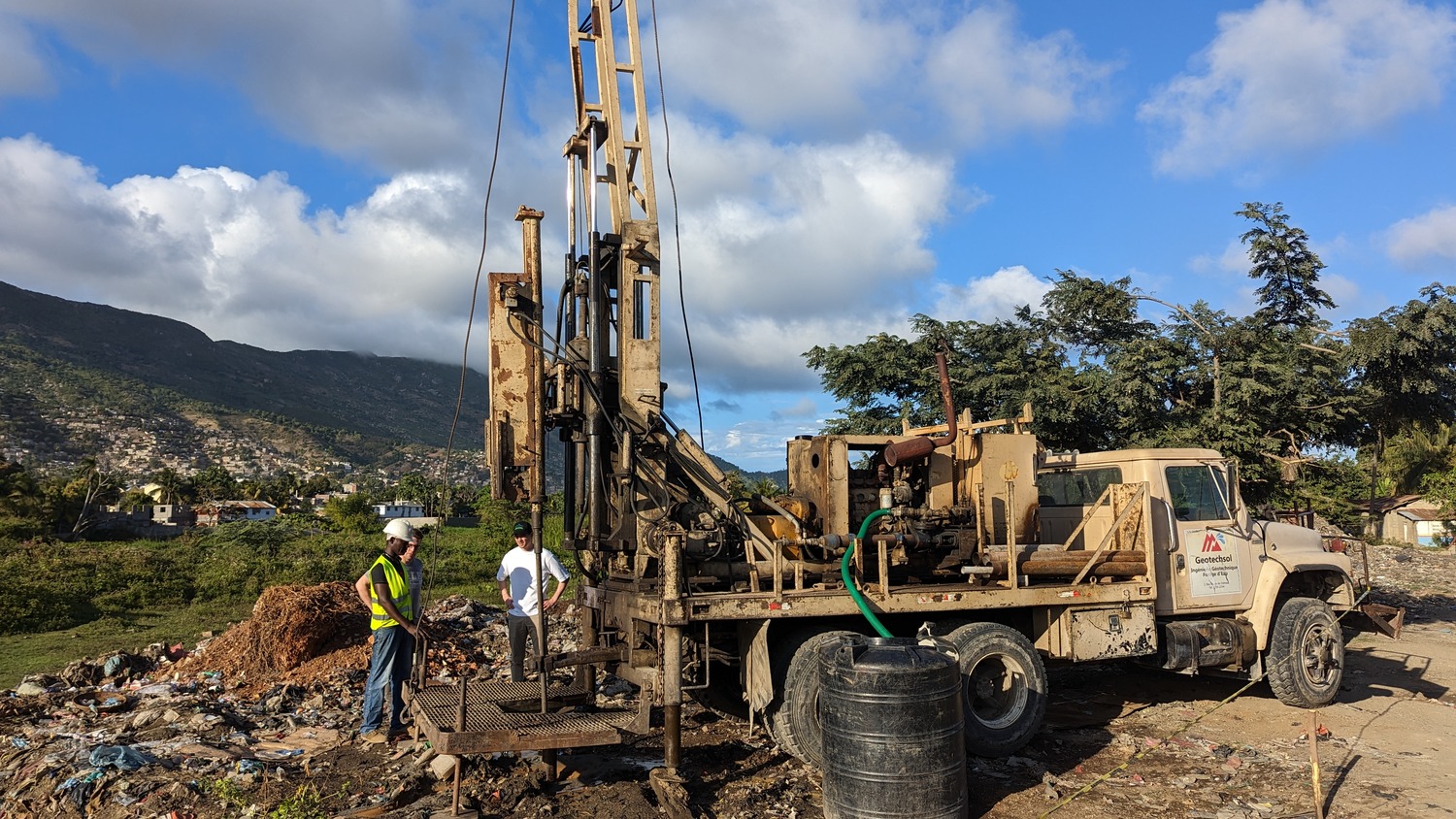 A drilling rig mounted on a truck is operating on a dirt site with two workers standing nearby; mountains and scattered buildings are visible in the background.