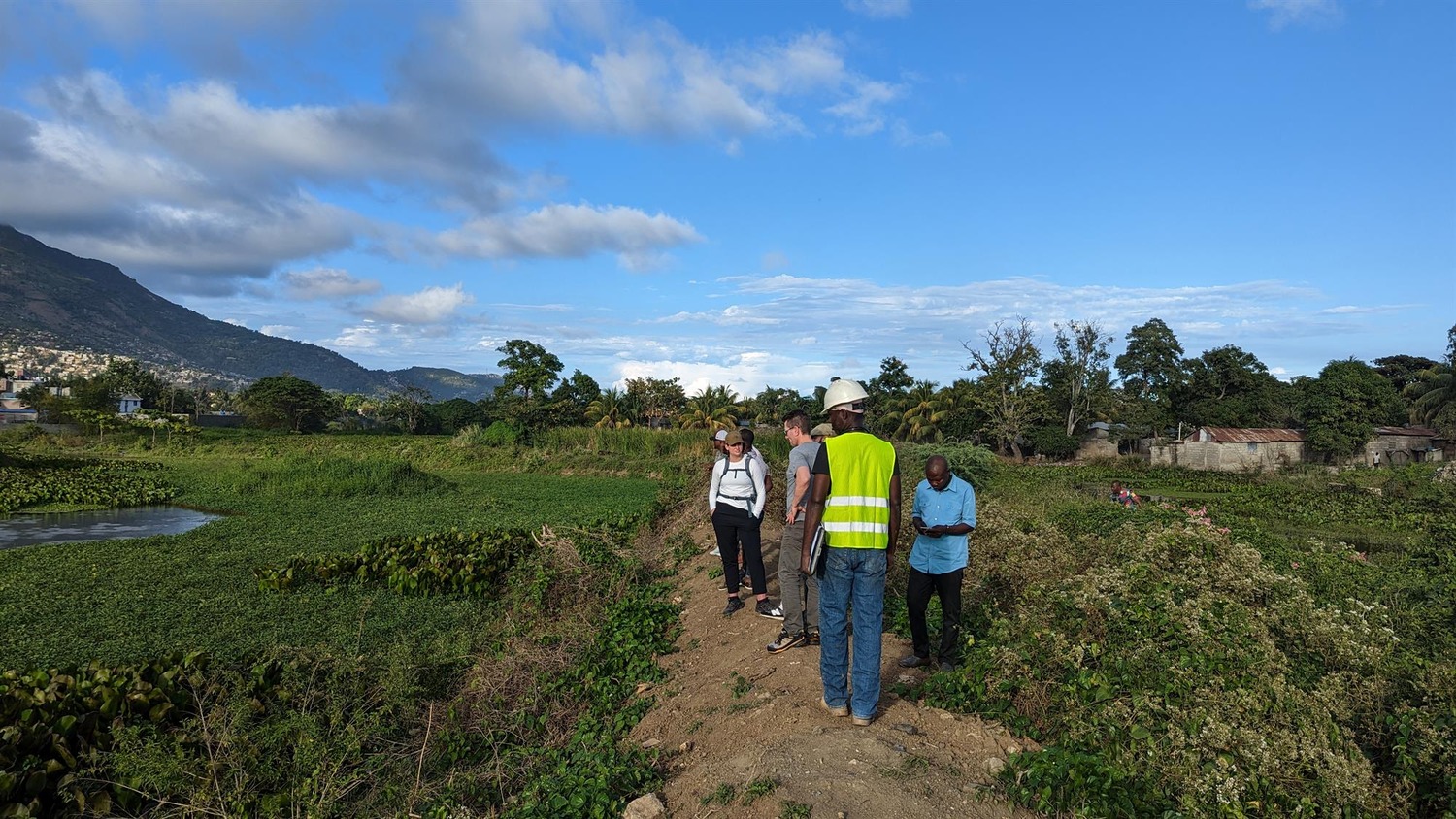 Several people, including one in a reflective vest and hard hat, stand on a dirt path in a green rural area with mountains and blue sky in the background.