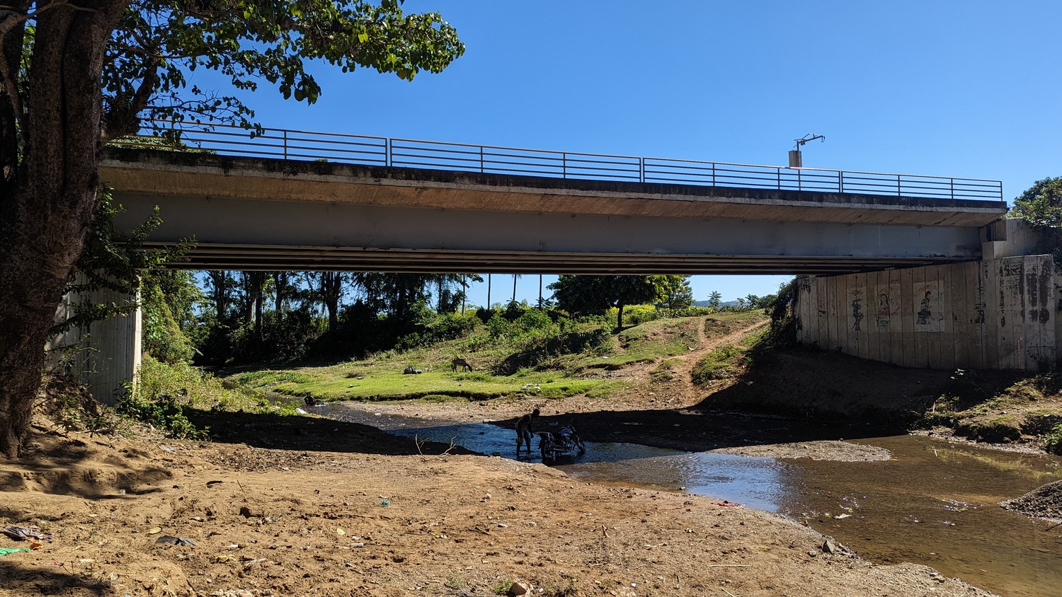 A concrete bridge crosses over a shallow creek with people standing in the water below on a sunny day. Trees and dirt paths are visible around the bridge.