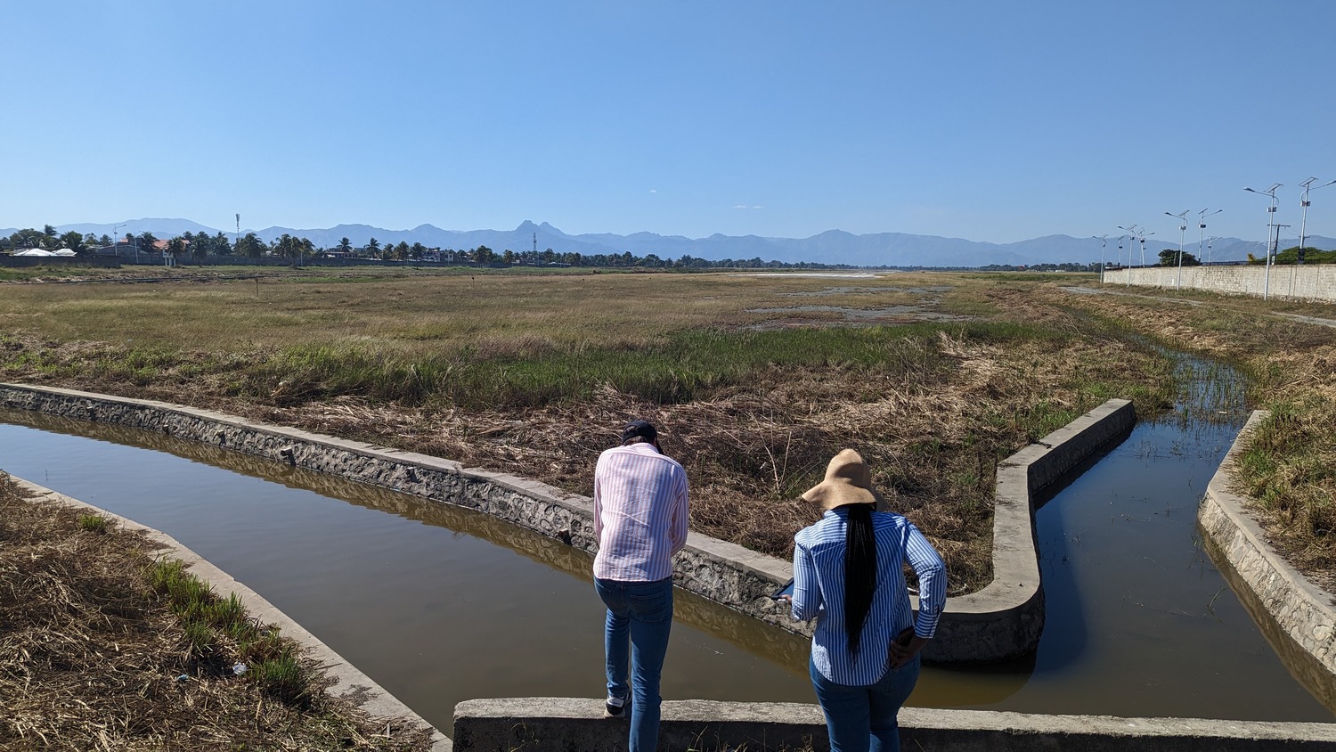 Two people wearing hats stand by a concrete irrigation canal, overlooking a dry field with mountains in the distance under a clear blue sky.