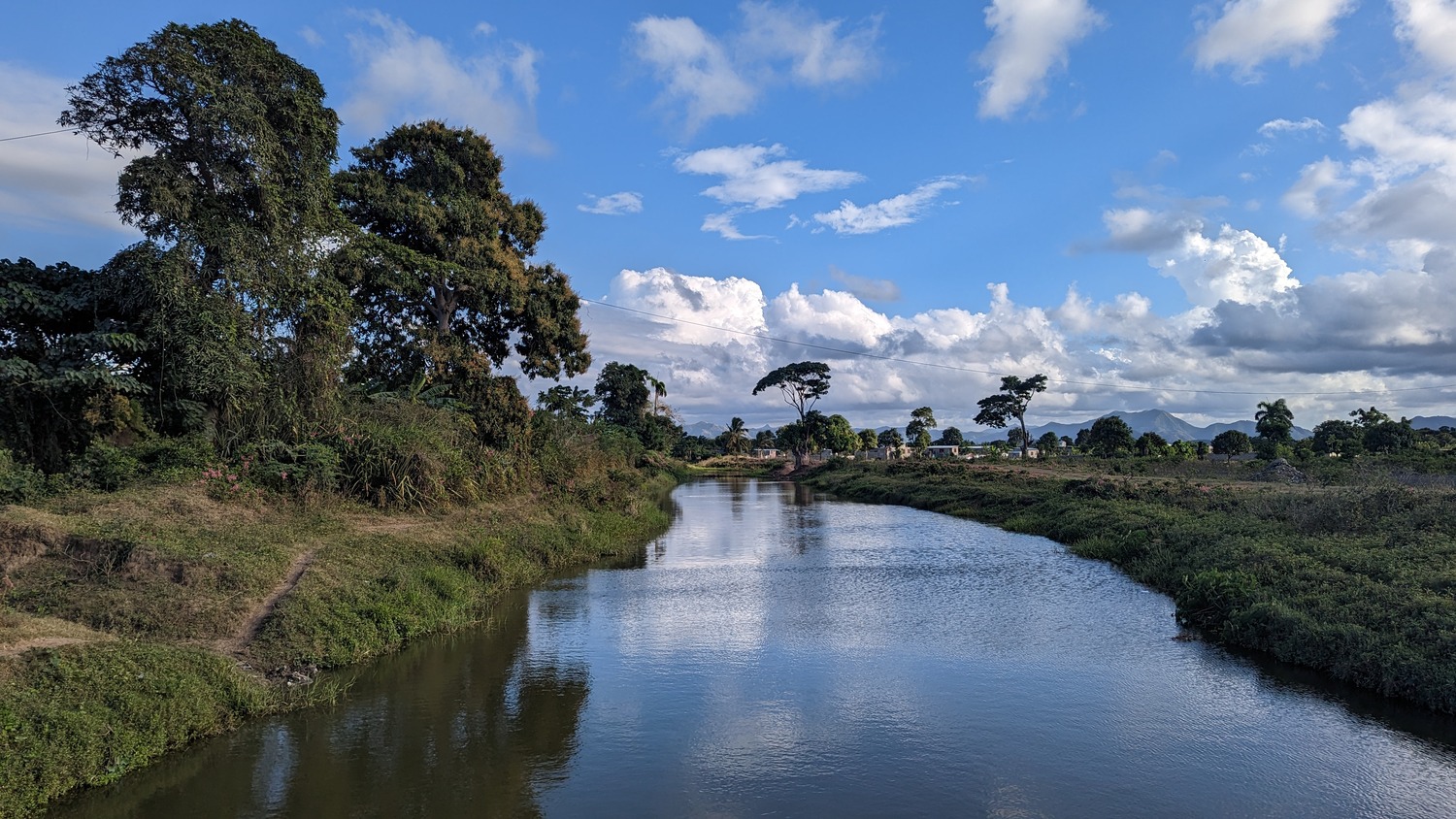 A calm river flows between grassy banks with trees on both sides under a blue sky filled with scattered clouds.