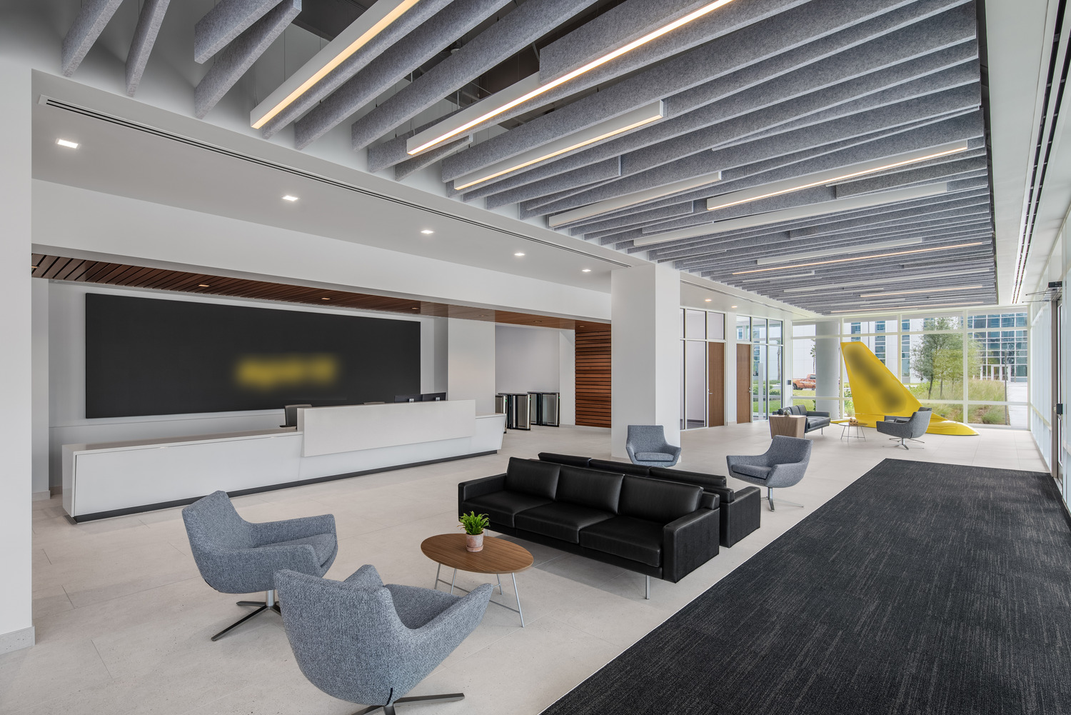 Modern office lobby with a black sofa, gray chairs, small round tables, and a large yellow sculpture by the window; reception desk visible in the background.