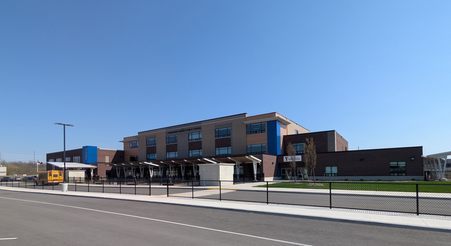 A modern, two-story school building with large windows, a blue accent section, and a yellow school bus parked outside under a clear blue sky.