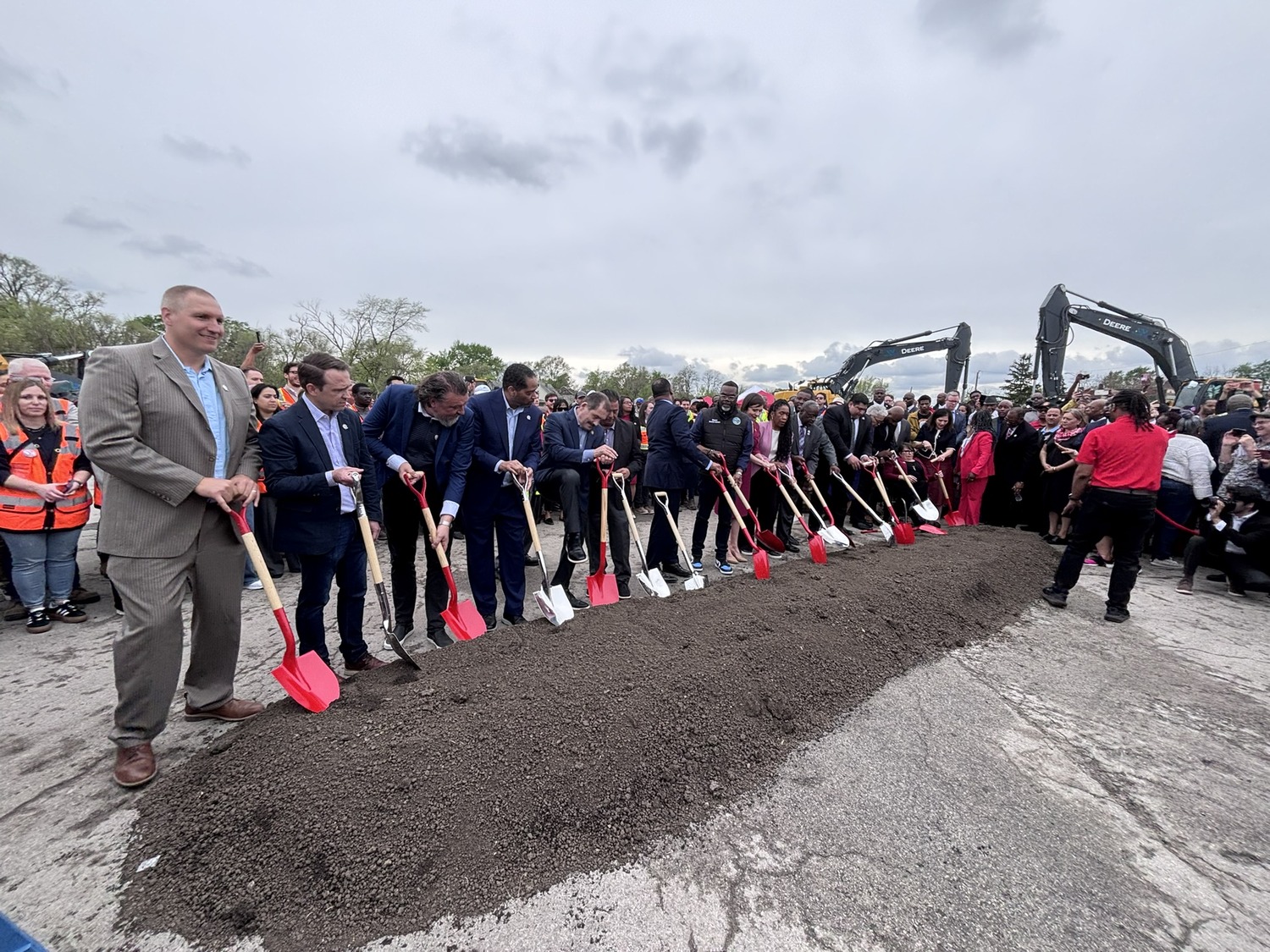A group of people in formal attire stand in a line, holding red shovels and breaking ground at a construction site, with excavators and a crowd in the background.