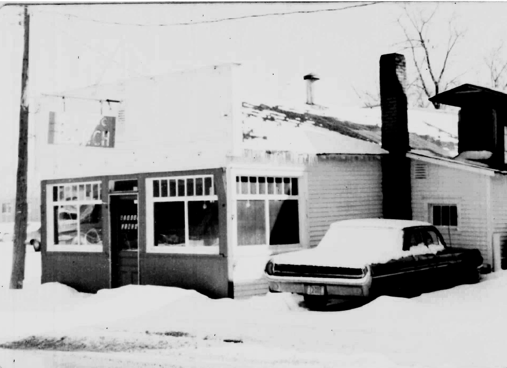 Black-and-white photo of a small building with large windows, snow on the ground and roof, and a classic car parked in front.