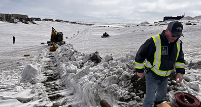 Workers in safety vests manage equipment and dig through snow in a large, snowy landscape with machinery and vehicles in the background.