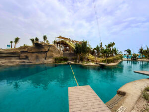 A scenic water park with clear blue pools, rock formations, palm trees, wooden bridges, and waterslides under a partly cloudy sky showcases exceptional entertainment infrastructure designed for relaxation and adventure.