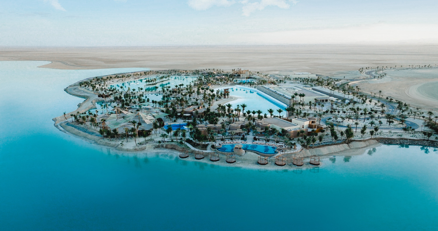 Aerial view of a resort island featuring pools, palm trees, and buildings surrounded by blue water and desert landscape, showcasing impressive entertainment infrastructure.