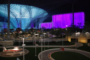 Indoor amusement park with illuminated glass dome, purple-lit building, miniature road track, streetlights, helicopter model, and trees—all showcasing impressive entertainment infrastructure under artificial lighting at night.