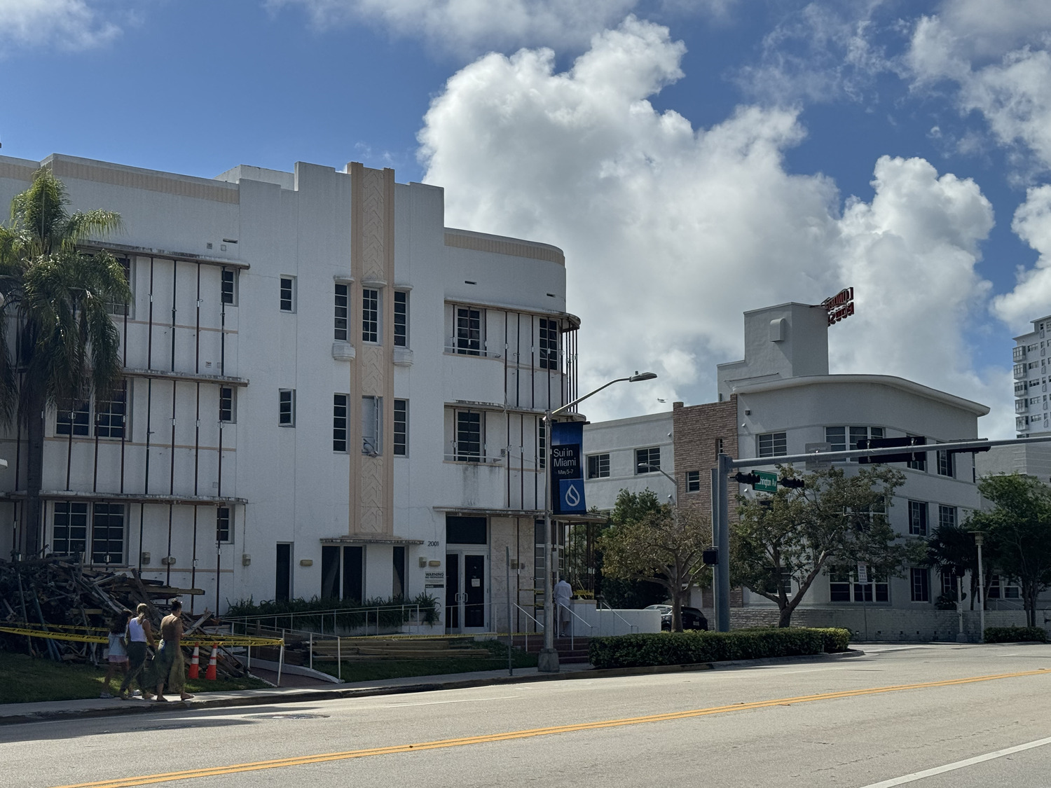A white art deco building in Miami-Dade County is under construction, with scaffolding on the facade. Orange cones and caution tape line the sidewalk as people walk nearby under a partly cloudy sky.