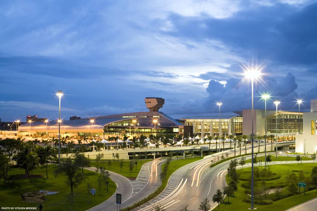 Modern airport terminal exterior at dusk with illuminated streetlights, car light trails on curved roads, palm trees, and a cloudy blue sky in the background.