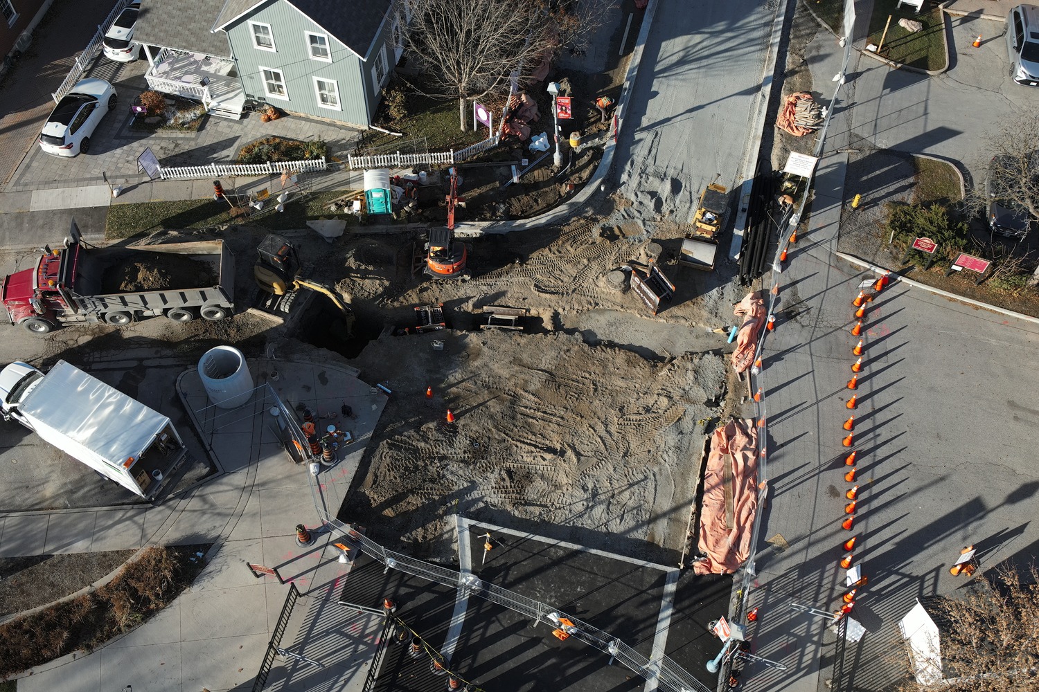 Aerial view of a road construction site with workers, vehicles, equipment, and a large trench beside a residential house. Traffic cones and barriers block off the area.