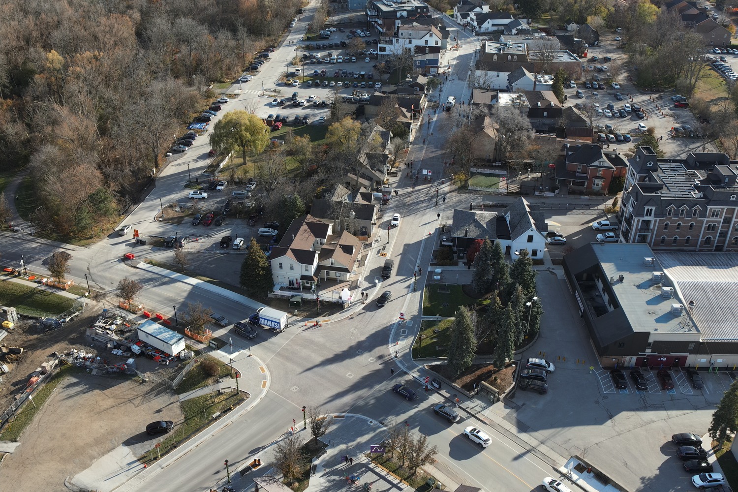 Aerial view of a suburban intersection with houses, parked cars, construction zones, trees, and nearby commercial buildings on a clear day.