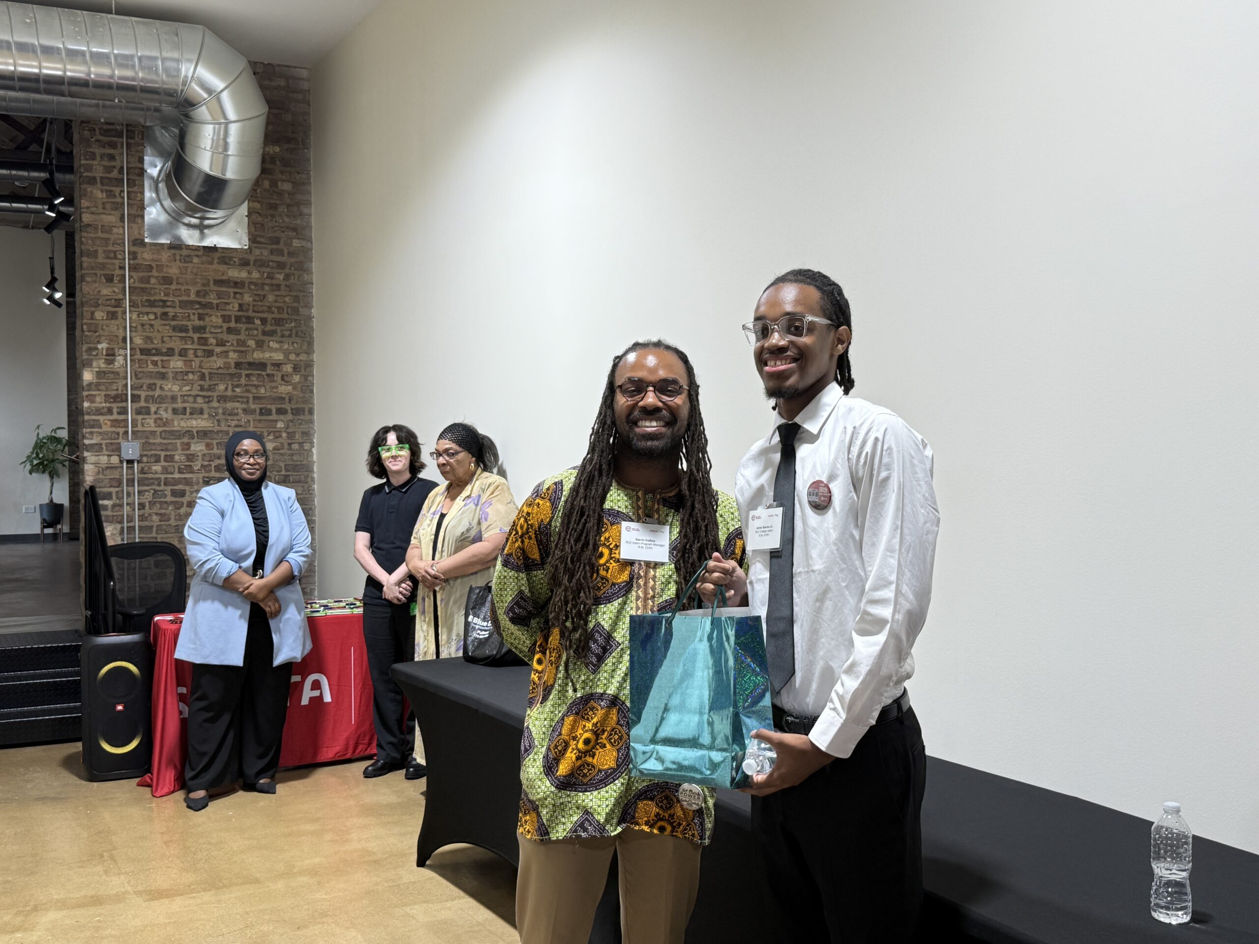 Two men stand together holding a blue gift bag, smiling at the camera. Three women stand in the background near a table covered with a red cloth.