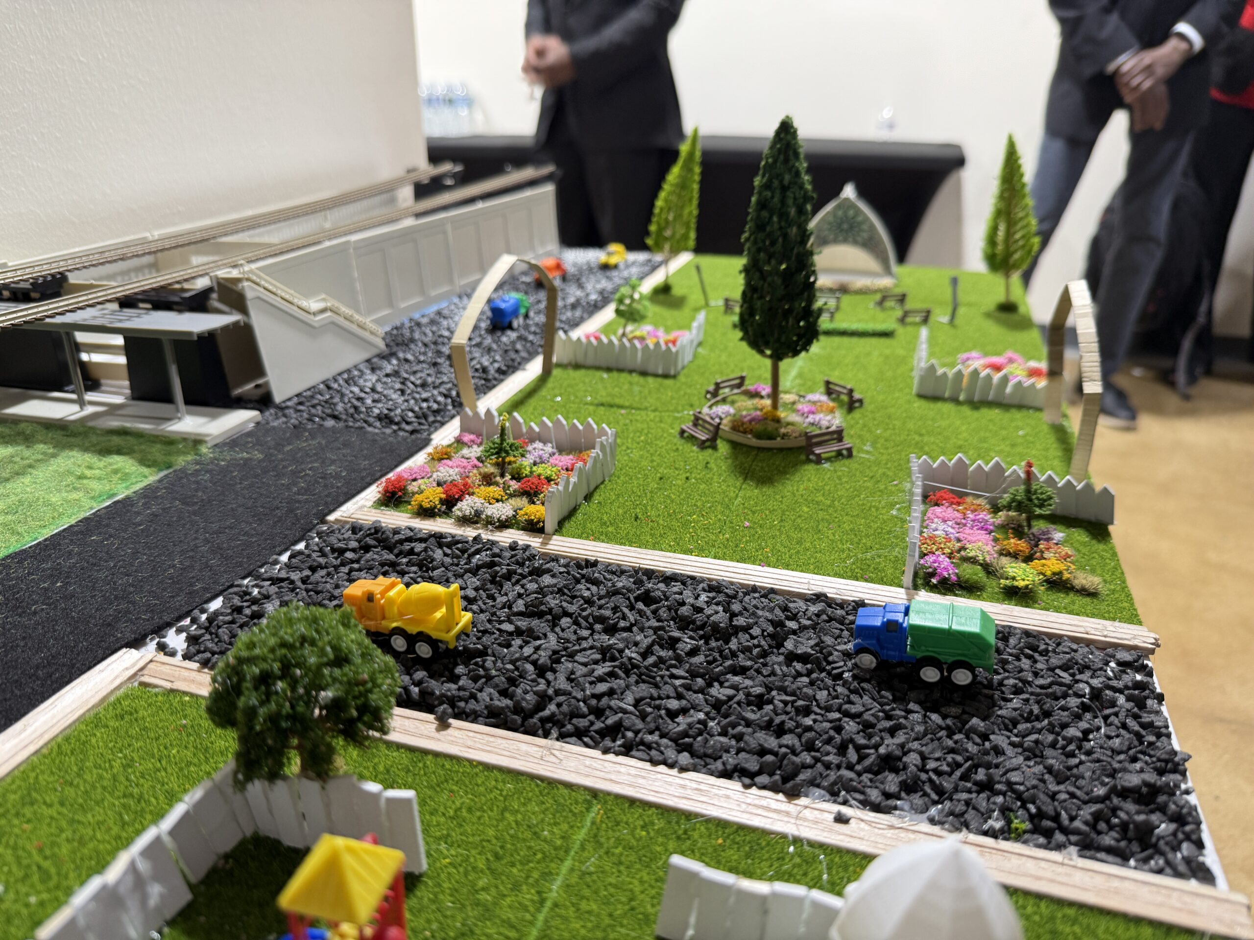 A close-up of a miniature model showing a park with trees, flowers, benches, a road with toy vehicles, and people standing in the background.
