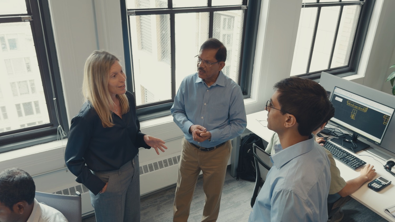 Three people stand and talk in an office near large windows, while a fourth person sits at a desk with a computer displaying data on the screen.