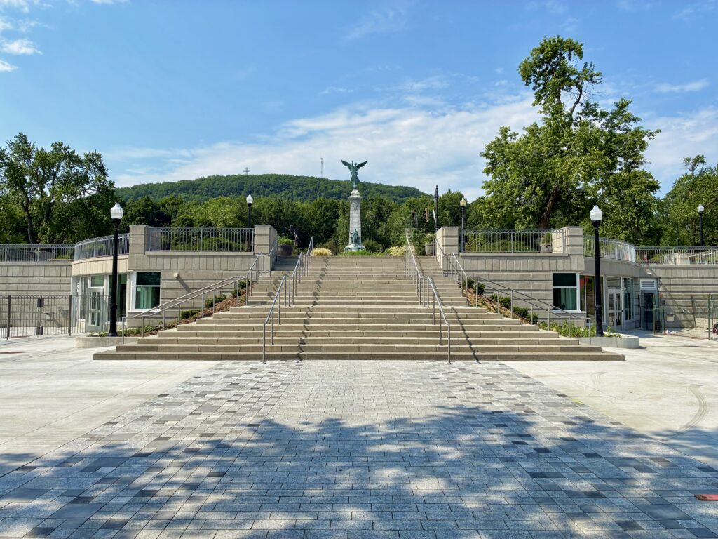 Wide stone staircase leads up to a tall monument with a winged statue, surrounded by greenery and trees under a clear sky.