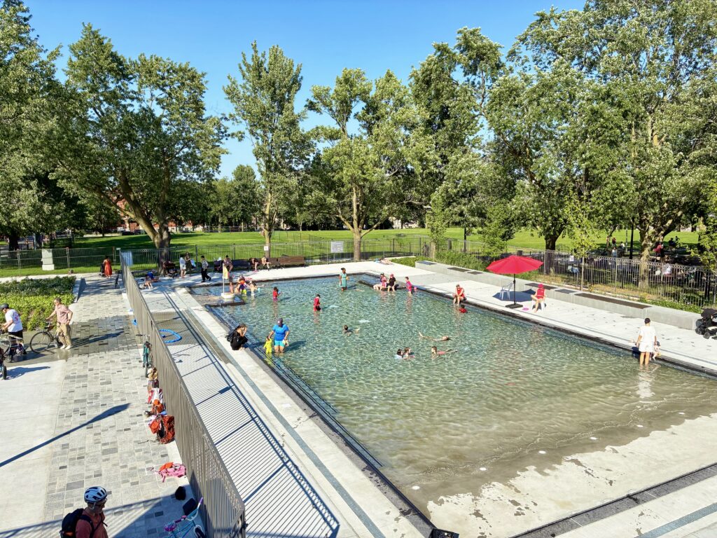 Children and adults swim and play in a shallow outdoor pool surrounded by trees and a park, with some people sitting or standing nearby.