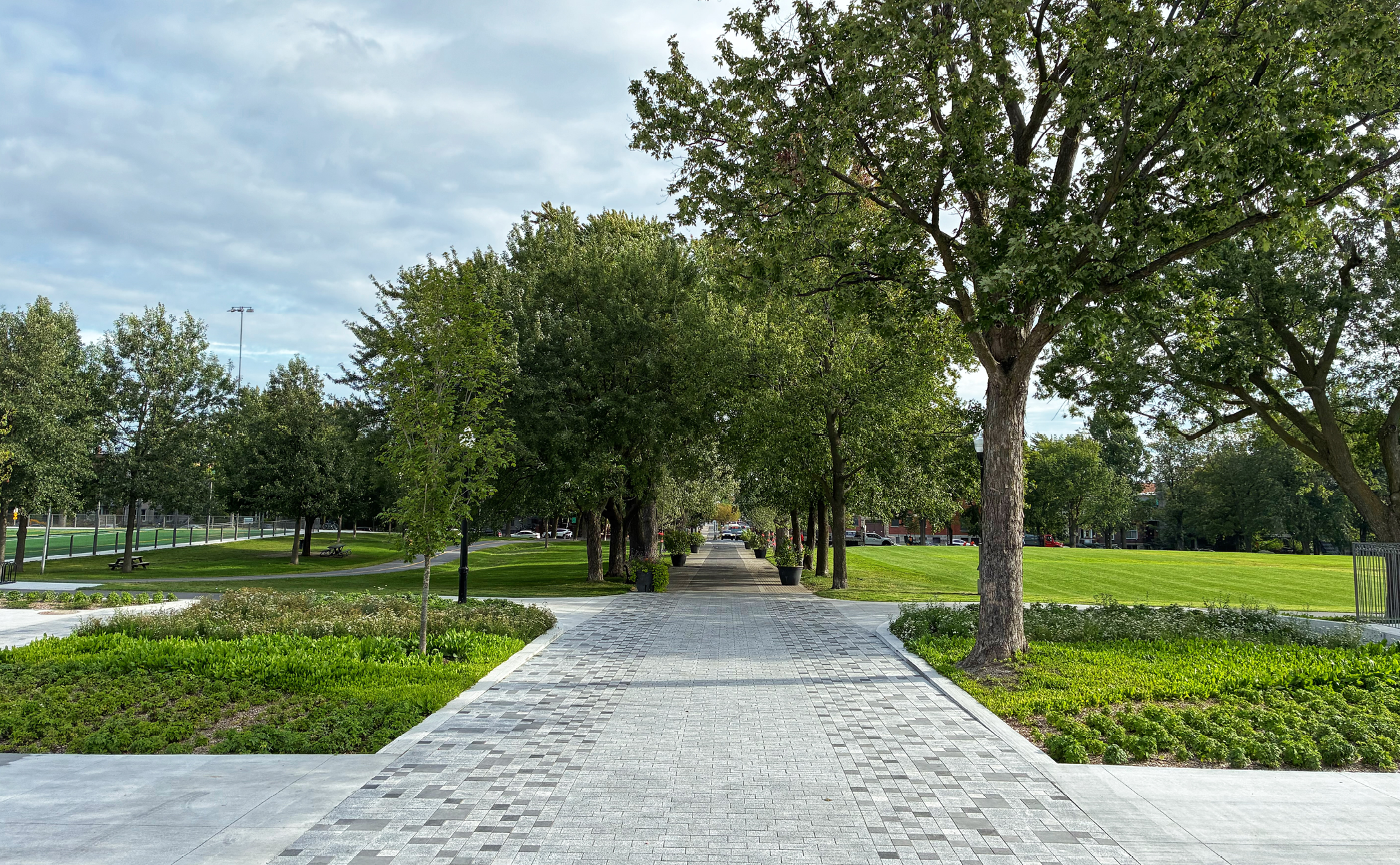 A paved walkway lined with trees runs through a park with green lawns on both sides under a cloudy sky.