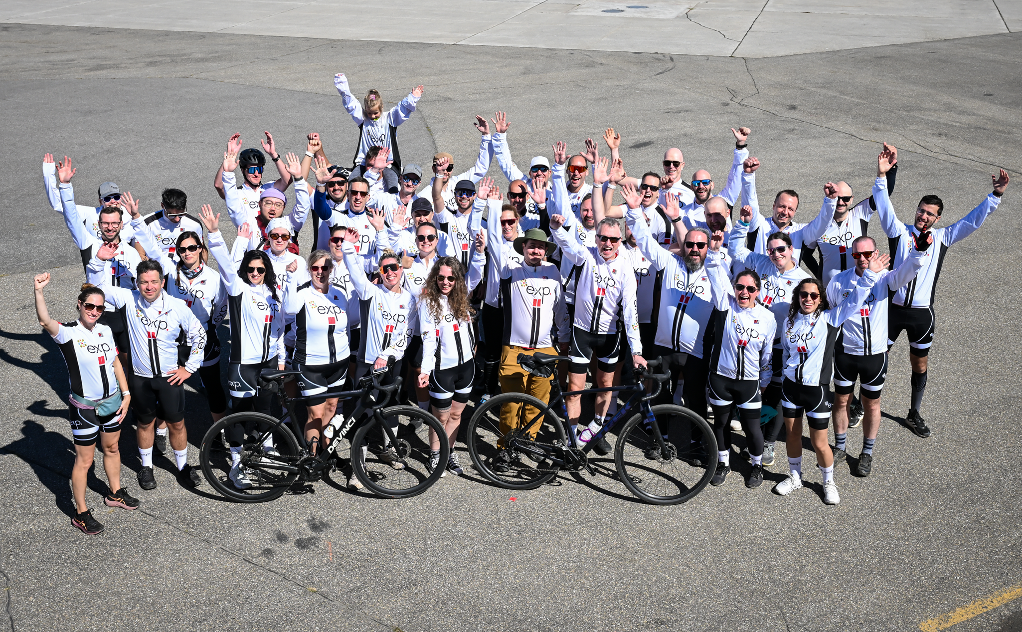 A large group of cyclists in matching white and black team jerseys poses for a photo on an asphalt surface with two bikes in the foreground.