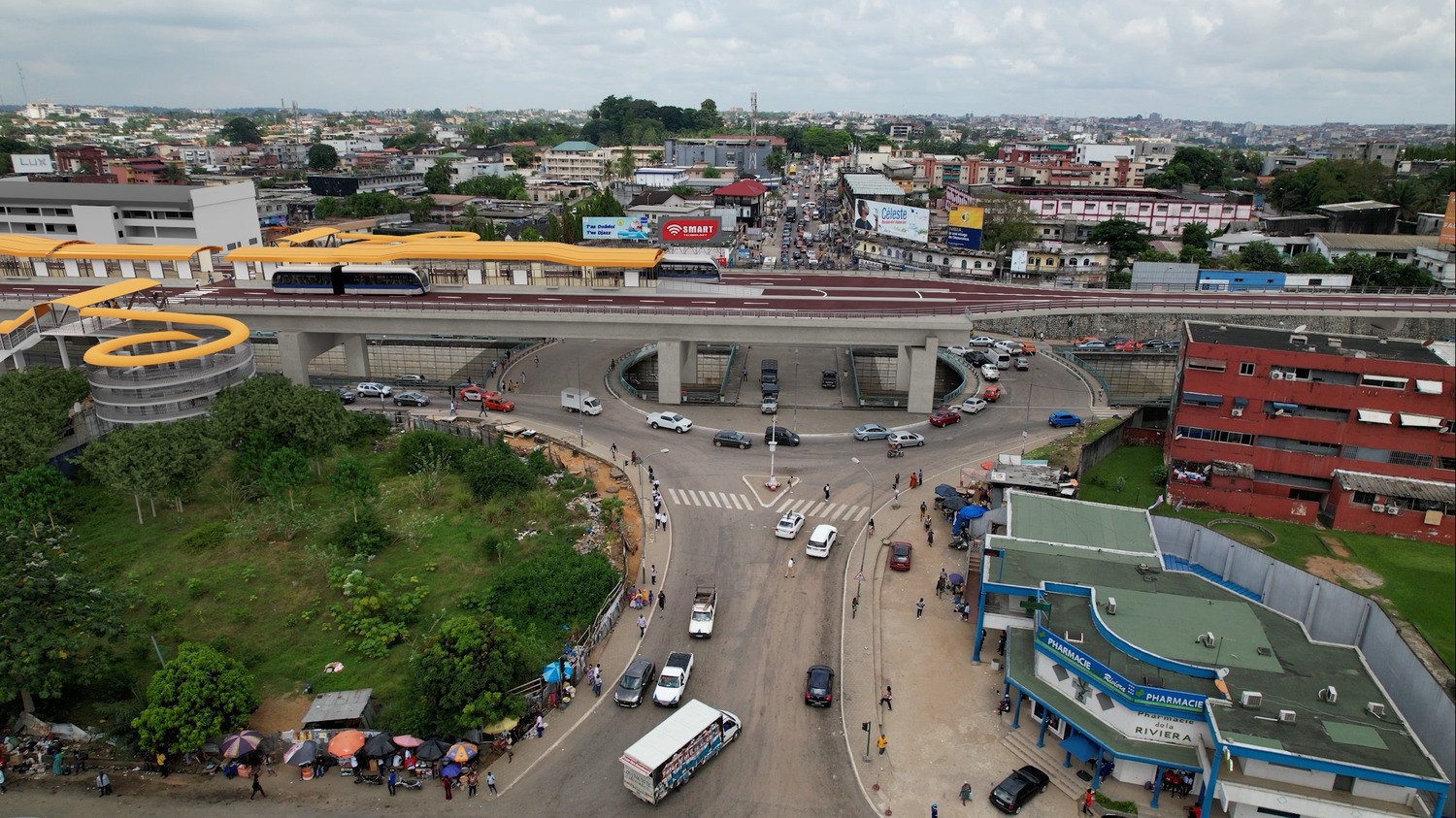 An elevated train track with a light rail vehicle runs above a busy city intersection, with surrounding streets, buildings, and a pharmacy visible below.