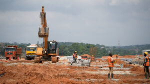 Construction workers in high-visibility vests work on a red-dirt site with an excavator and dump trucks.