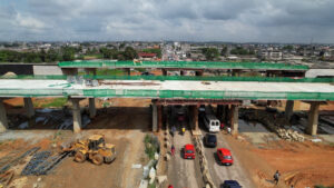 Aerial view of an unfinished elevated highway bridge under construction above a busy road with cars and a yellow construction vehicle.
