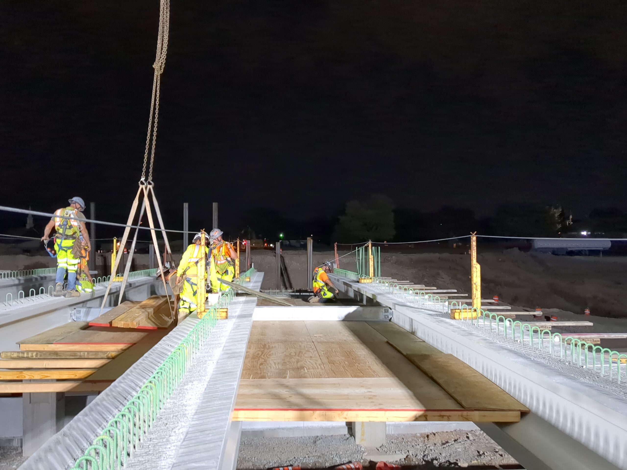 Construction workers in high-visibility vests and hard hats work on an elevated bridge structure at night.