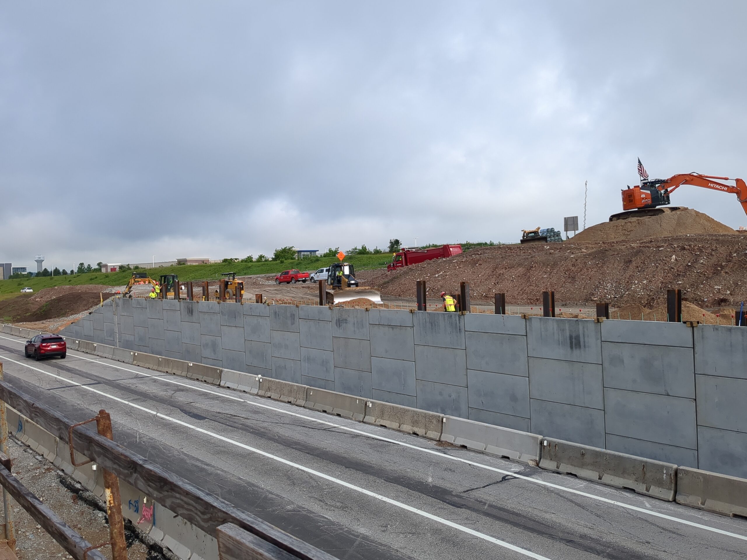 A highway construction site featuring a concrete retaining wall, various pieces of heavy machinery, and a red car driving in the foreground.