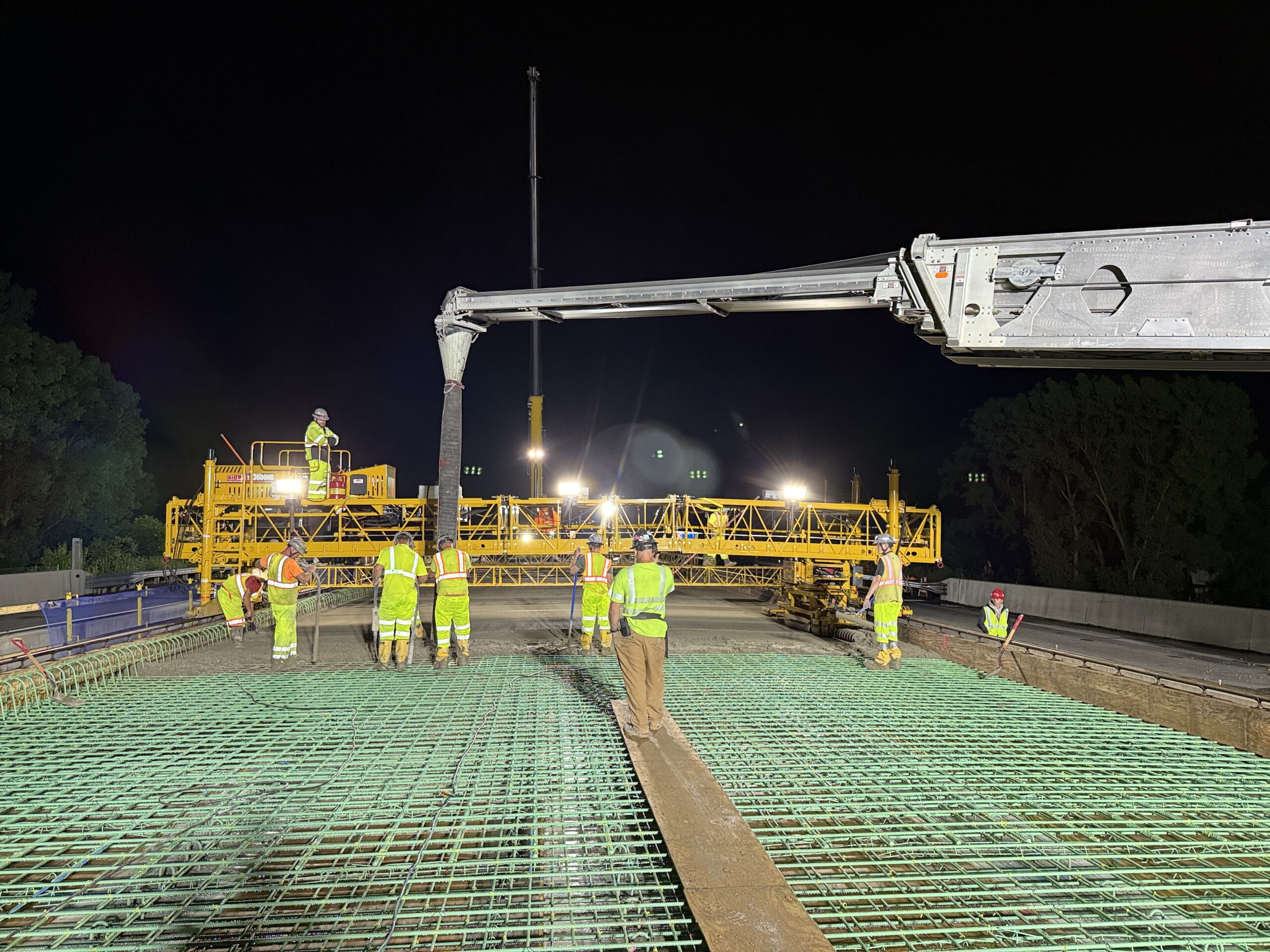 Construction workers in high-visibility gear work on a bridge deck with green rebar at night, as a concrete pump truck pours material.